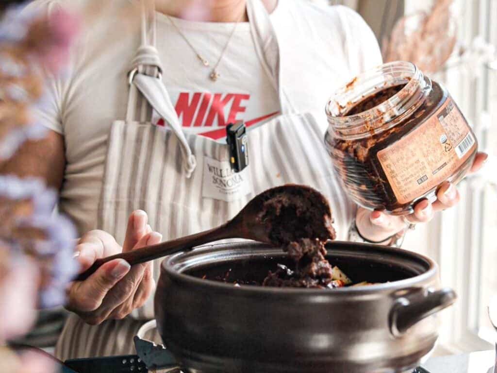 A person in a striped apron is holding a jar of doenjang, scooping it with a wooden spoon into a pot. They wear a white shirt with a red Nike logo. The scene is set in a kitchen with soft lighting.